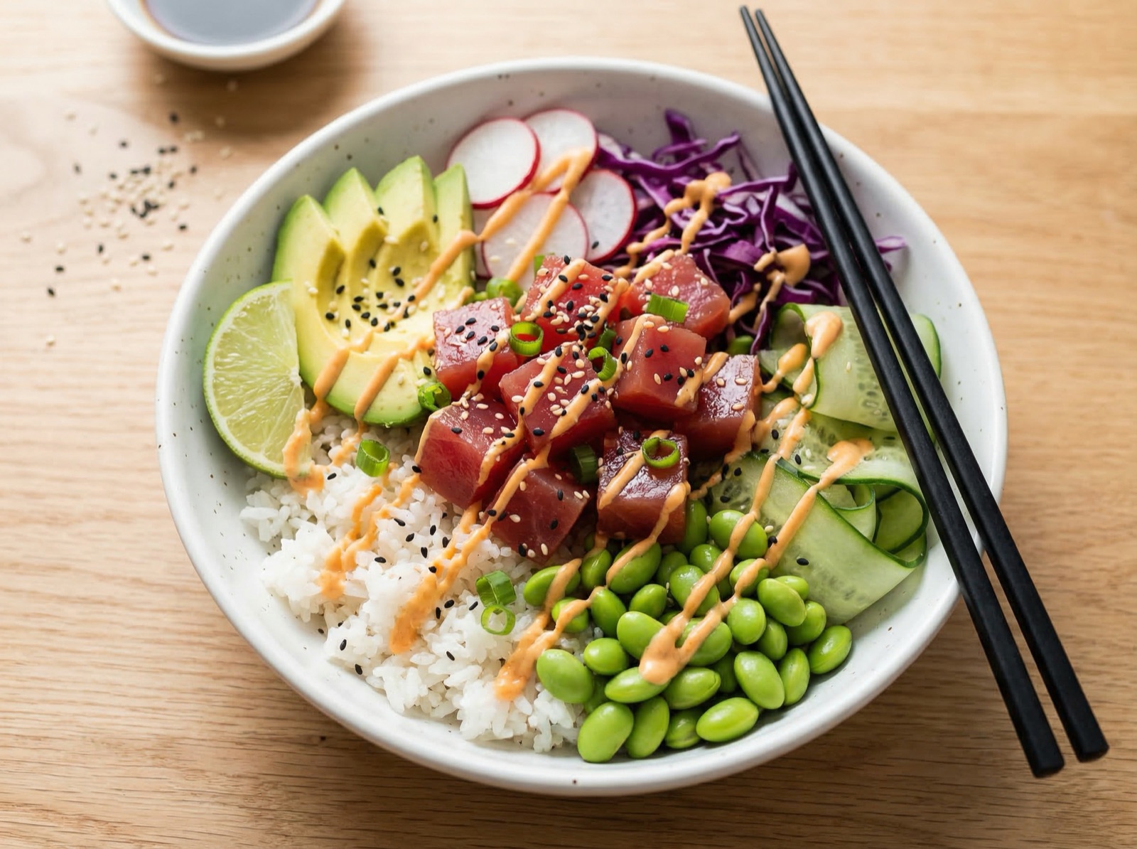Overhead close-up of a vibrant poke bowl with sushi-grade tuna, rice, avocado, edamame, and spicy mayo drizzle on a light oak table